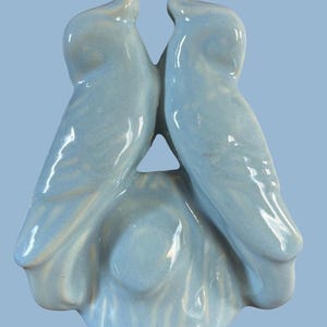 May include: A light blue ceramic sculpture of two stylized birds facing each other. The birds are connected at the base and have a glossy finish. The sculpture is on a light blue background.