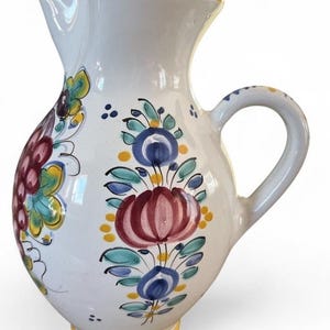 Puede incluir: Jarra de cerámica blanca con borde y base amarillos, adornada con diseños florales pintados a mano en rojo, azul, verde y amarillo. La jarra tiene un asa curva y un pico vertedor.