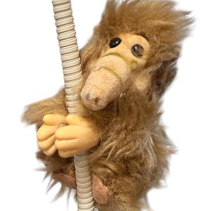 Puede incluir: Un peluche de extraterrestre marrón y peludo, con grandes ojos negros y un hocico beige. El extraterrestre se aferra a un poste crema y estriado con sus manos beige. El juguete tiene una apariencia de dibujos animados.