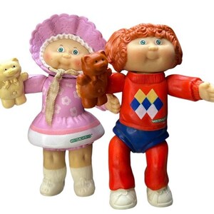 May include: Two Cabbage Patch Kids dolls, one wearing a pink dress and a white bonnet, the other wearing a red sweater with a diamond pattern and blue pants. Both dolls are holding teddy bears.