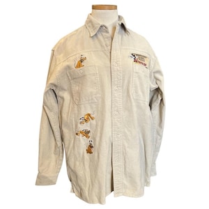Puede incluir: Camisa beige con botones con diseños bordados del personaje de dibujos animados Pluto. La camisa tiene dos bolsillos en el pecho y mangas largas. El texto "Walt Disney World Movie Magic" está bordado en el pecho derecho.