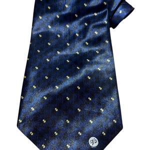 Puede incluir: Una corbata de seda azul oscuro con un pequeño patrón de lunares blancos y dorados. La corbata tiene una pequeña etiqueta blanca con la letra "V" y el número "2" en ella.