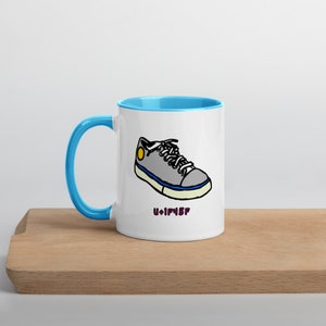 Könnte beinhalten: Eine weiße Keramiktasse mit hellblauem Rand und Henkel. Die Tasse zeigt eine Comiczeichnung eines grauen Sneakers mit gelbem Akzent und dem Text "U+1F45F".