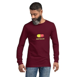 May include: A maroon long-sleeve shirt featuring a graphic of a yellow and red capsule with the text "100 mg SERTRALINE" in yellow. The shirt is worn by a person with a beard and blue jeans.