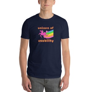 以下が含まれることがあります： 虹の上を走るピンクのユニコーンが描かれたネイビーブルーのTシャツ。オレンジ色の「unicorn of usability」という文字が書かれています。