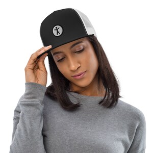 Puede incluir: Una gorra de camionero negra y blanca con un logotipo blanco de una figura de palo en un círculo. La gorra la lleva puesta una persona con el pelo largo y negro.