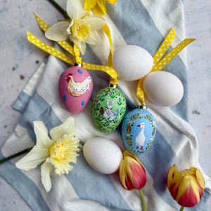 May include: A collection of Easter decorations featuring painted eggs with animal designs, white eggs, daffodils, and tulips. The eggs are decorated with ribbons and arranged on a blue and white striped cloth.
