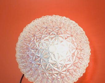May include: A clear, spherical lamp with a textured, starburst pattern. The lamp is lit, emitting a bright glow. A black cord extends from the bottom of the lamp. The background is a solid orange-red.