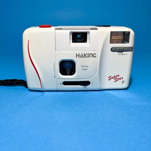 May include: White Haking Super Star F 35mm film camera with a red accent line and a black wrist strap. The camera has the text "Haking Focus Free" and "Flash >".
