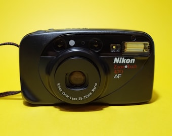 May include: Black Nikon Zoom Touch 470 AF camera with a 35-70mm macro lens. The camera has a built-in flash and a red and black wrist strap. The camera is on a yellow background.