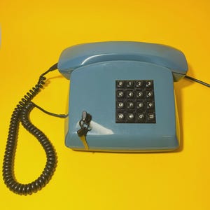 May include: A vintage blue telephone with a keypad and a coiled handset cord. The phone has a lock and key. The keypad features numbers and letters. The phone is set against a bright yellow background.