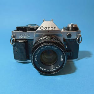 Könnte beinhalten: Eine Vintage Canon AE-1 Kamera mit einem 50mm 1:1.8 Objektiv. Das Kameragehäuse ist silber und schwarz, mit dem Canon-Logo. Das Objektiv ist mit "CANON LENS FD 50mm 1:1.8" und "LENS MADE IN JAPAN" beschriftet.