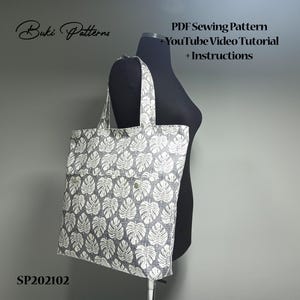 May include: A grey tote bag with a white leaf pattern. The bag has a large pocket on the front. The bag is on a black dress form. The text "Buki Patterns" is in the top left corner of the image. The text "PDF Sewing Pattern", "YouTube Video Tutorial", and "Instructions" is in the top right corner of the image. The text "SP202102" is in the bottom left corner of the image.