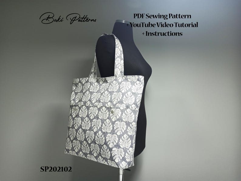 May include: A grey tote bag with a white leaf pattern. The bag has a large pocket on the front. The bag is on a black dress form. The text "Buki Patterns" is in the top left corner of the image. The text "PDF Sewing Pattern", "YouTube Video Tutorial", and "Instructions" is in the top right corner of the image. The text "SP202102" is in the bottom left corner of the image.