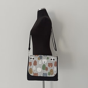 May include: A black crossbody bag with a gray and white fabric flap featuring a repeating pattern of bears, foxes, and trees. The bag has a black adjustable strap.