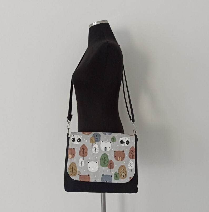 May include: A black crossbody bag with a gray and white fabric flap featuring a repeating pattern of bears, foxes, and trees. The bag has a black adjustable strap.