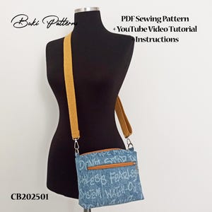 May include: A denim crossbody bag with a brown leather zip pull and strap. The bag features white graffiti-style text and is displayed on a black mannequin. The text on the bag reads "DON'T STOP FEARLESS SYSTEM WATCH-ON".