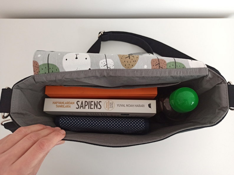 May include: A black canvas messenger bag with a gray lining. The bag is open and contains a book titled "Sapiens", a water bottle, and a blue yoga mat.