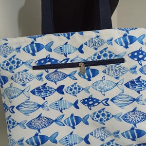 May include: A white canvas tote bag with blue fish designs. The bag has a zippered pocket on the front and two navy blue handles.