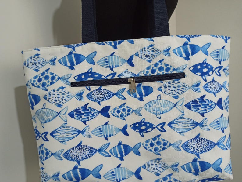 May include: A white canvas tote bag with blue fish designs. The bag has a zippered pocket on the front and two navy blue handles.