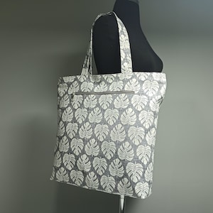 May include: A grey and white tote bag with a tropical leaf pattern. The bag has a zipped pocket on the front.