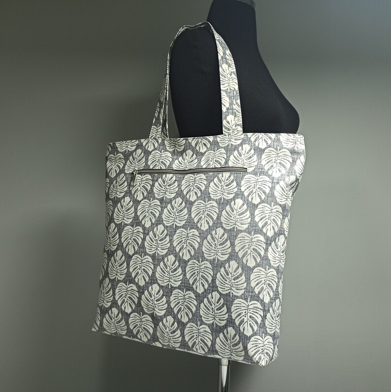 May include: A grey and white tote bag with a tropical leaf pattern. The bag has a zipped pocket on the front.
