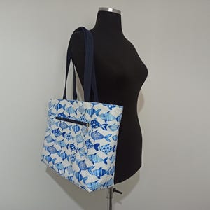 May include: A white tote bag with a blue fish pattern. The bag has a zippered pocket on the front and two navy blue straps.