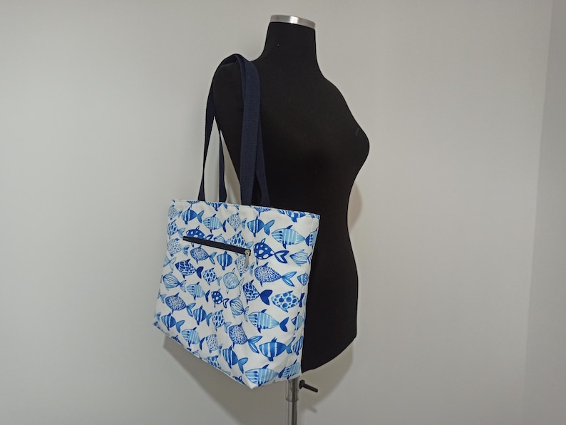 May include: A white tote bag with a blue fish pattern. The bag has a zippered pocket on the front and two navy blue straps.
