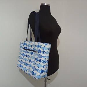 May include: A white tote bag with a blue fish pattern. The bag has a zippered pocket on the front and two navy blue straps.