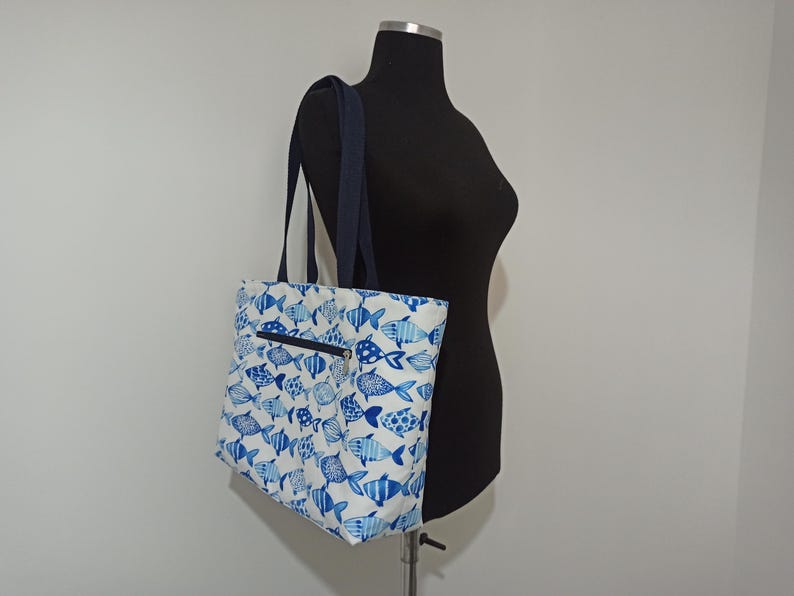 May include: A white tote bag with a blue fish pattern. The bag has a zippered pocket on the front and two navy blue straps.