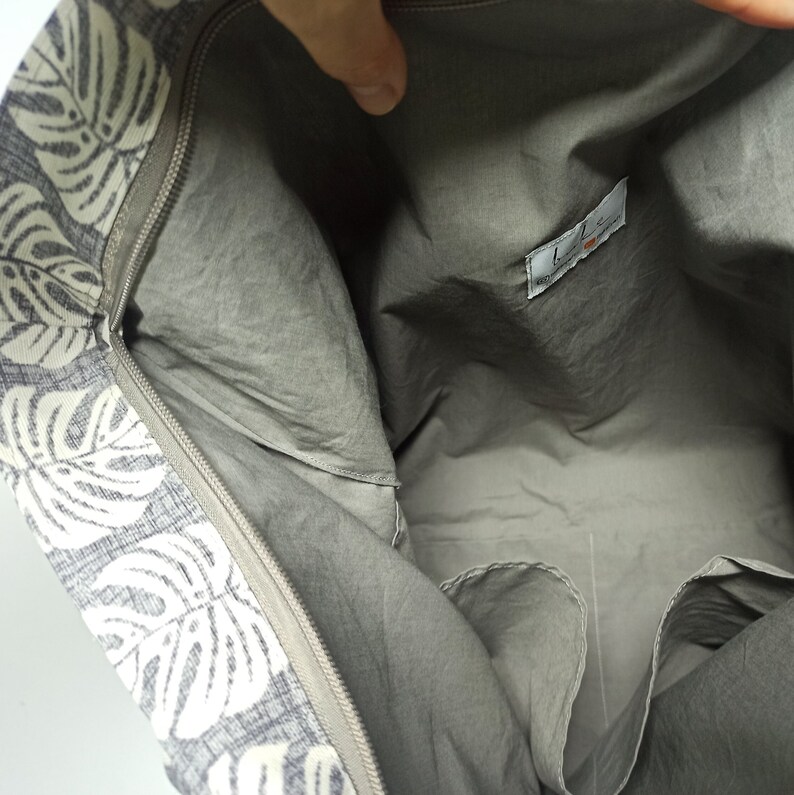 May include: The inside of a grey tote bag with a white and grey floral print. The bag has a zipper closure and a small pocket on the inside.