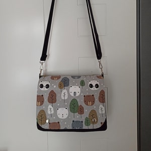 May include: A gray and white shoulder bag with a black strap. The bag has a print of bears, trees, and other woodland creatures.