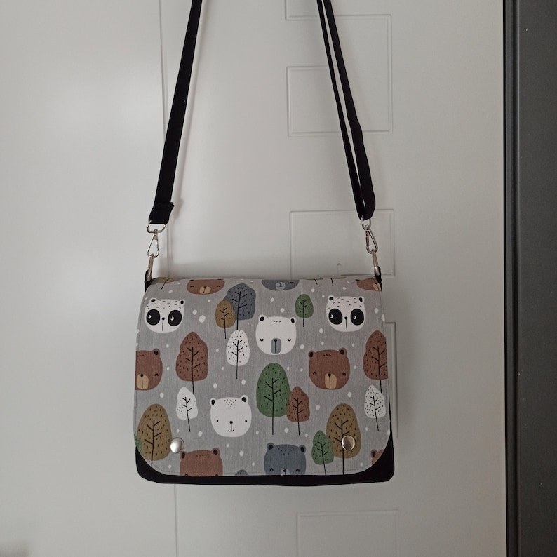 May include: A gray and white shoulder bag with a black strap. The bag has a print of bears, trees, and other woodland creatures.