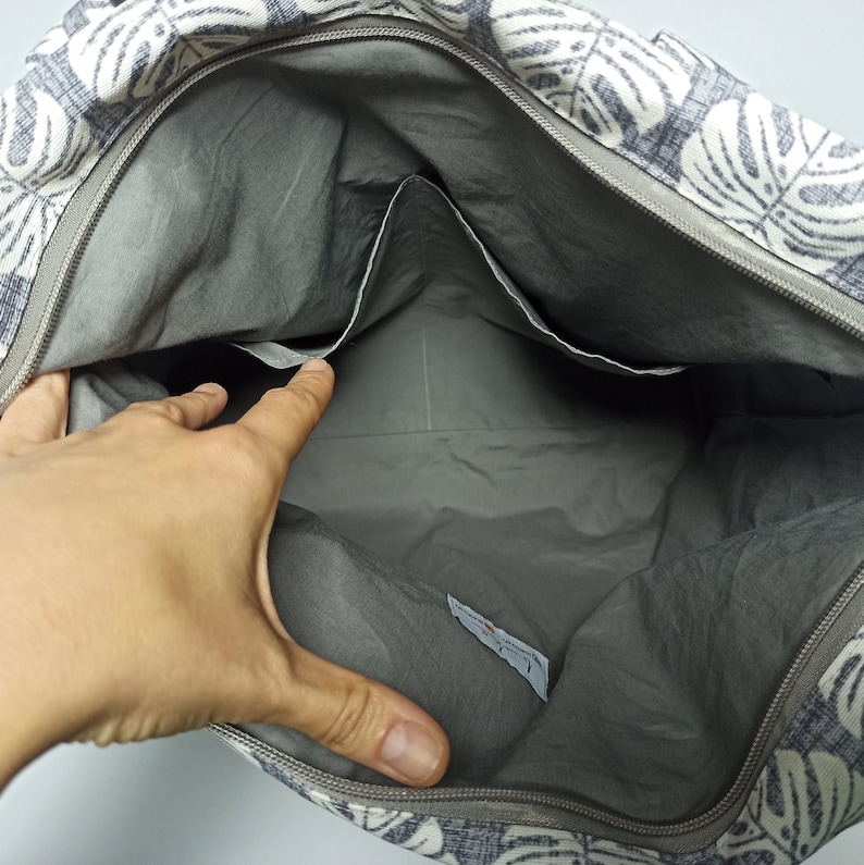 May include: A grey canvas tote bag with a white and grey leaf pattern. The bag is open and the inside is lined with grey fabric. There are two pockets on the inside of the bag.