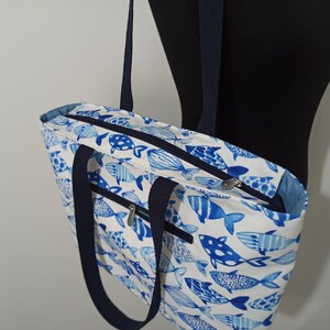 May include: A white tote bag with blue fish designs. The bag has two navy blue handles and a zipper closure.