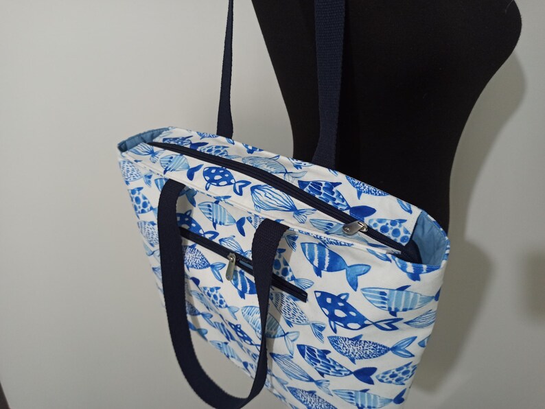 May include: A white tote bag with blue fish designs. The bag has two navy blue handles and a zipper closure.
