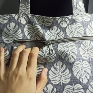 May include: A grey canvas tote bag with a white monstera leaf pattern. The bag has a zipped pocket and a silver zipper pull.