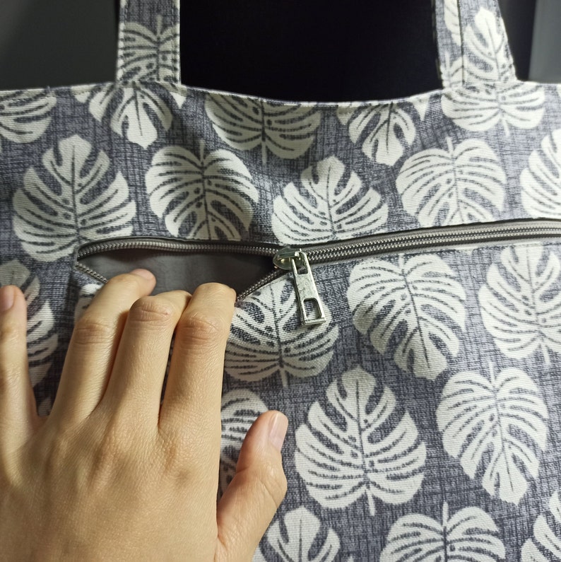 May include: A grey canvas tote bag with a white monstera leaf pattern. The bag has a zipped pocket and a silver zipper pull.