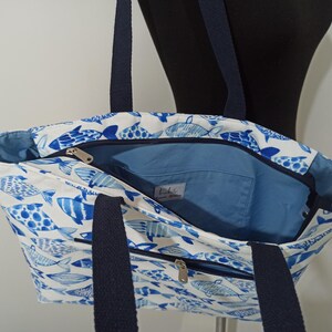 May include: A white canvas tote bag with a blue and white fish pattern. The bag has two navy blue straps and a zippered pocket on the front.