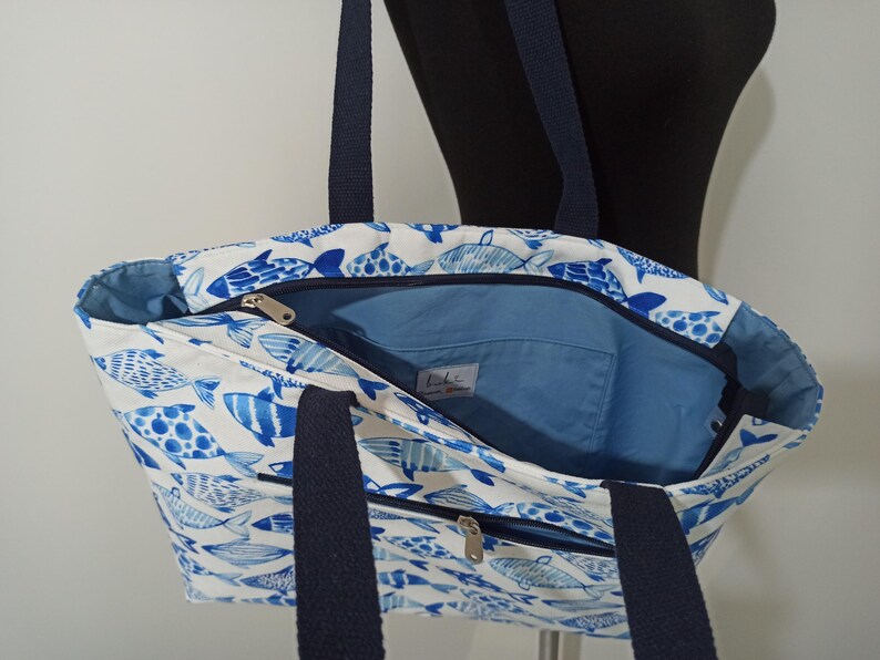 May include: A white canvas tote bag with a blue and white fish pattern. The bag has two navy blue straps and a zippered pocket on the front.