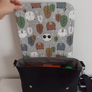 May include: A black canvas messenger bag with a gray and white fabric lining featuring a repeating pattern of bears, pandas, and leaves. The bag has a flap closure with two silver snaps and a long adjustable strap.