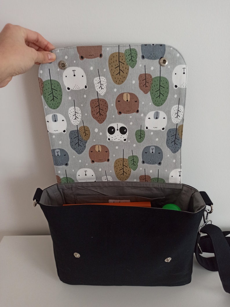 May include: A black canvas messenger bag with a gray and white fabric lining featuring a repeating pattern of bears, pandas, and leaves. The bag has a flap closure with two silver snaps and a long adjustable strap.