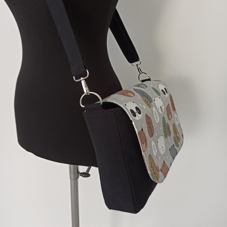 May include: A black and gray fabric crossbody bag with a flap closure. The flap is decorated with a white and gray print of cartoon bears and trees.