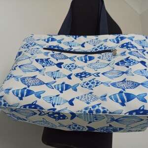 May include: A white tote bag with a blue and white fish pattern. The bag has a zippered pocket on the front and two navy blue straps.