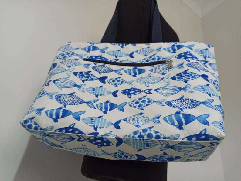 May include: A white tote bag with a blue and white fish pattern. The bag has a zippered pocket on the front and two navy blue straps.