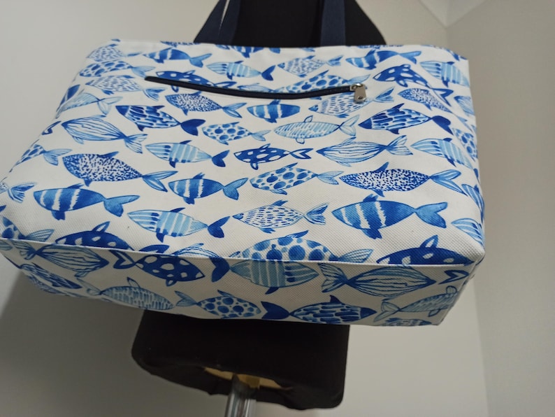 May include: A white canvas tote bag with a blue and white fish pattern. The bag has a zippered pocket on the front.