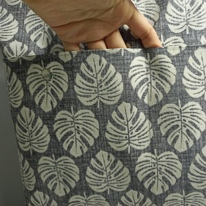 May include: A grey canvas tote bag with a white monstera leaf pattern. The bag has a zipped pocket on the front.