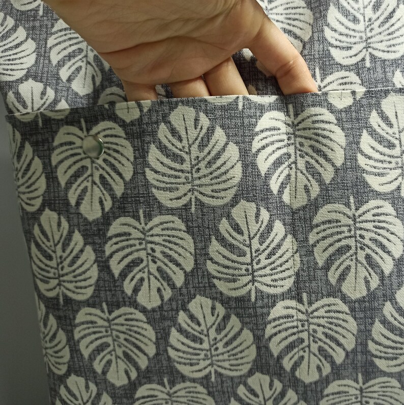 May include: A grey canvas tote bag with a white monstera leaf pattern. The bag has a zipped pocket on the front.