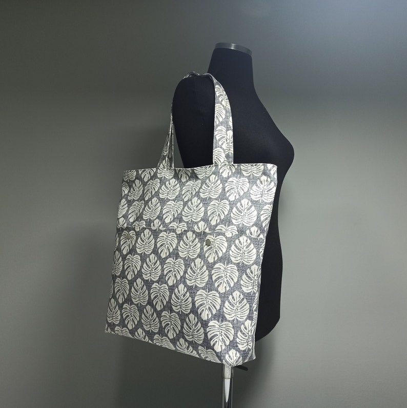 May include: A grey and white tote bag with a leaf pattern. The bag has a large pocket on the front.