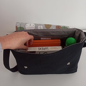 May include: A black canvas messenger bag with a gray lining. The bag is open and contains a book titled "Sapiens", a green water bottle, and a few other items.